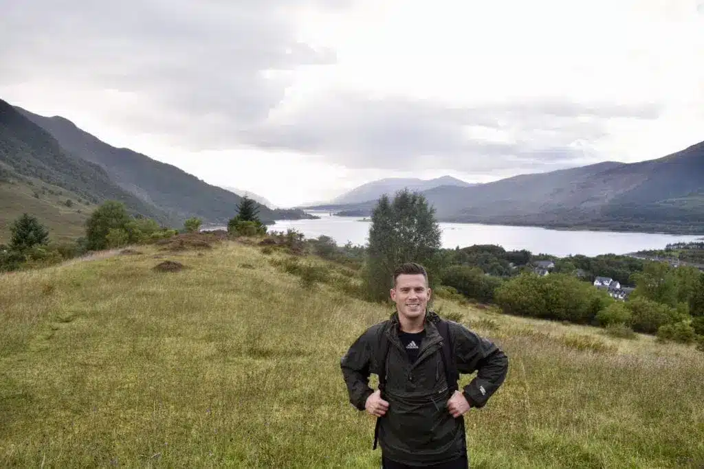 Kristoffer Fons, do Destination Well Known, numa trilha gramada com um lago e montanhas ao fundo