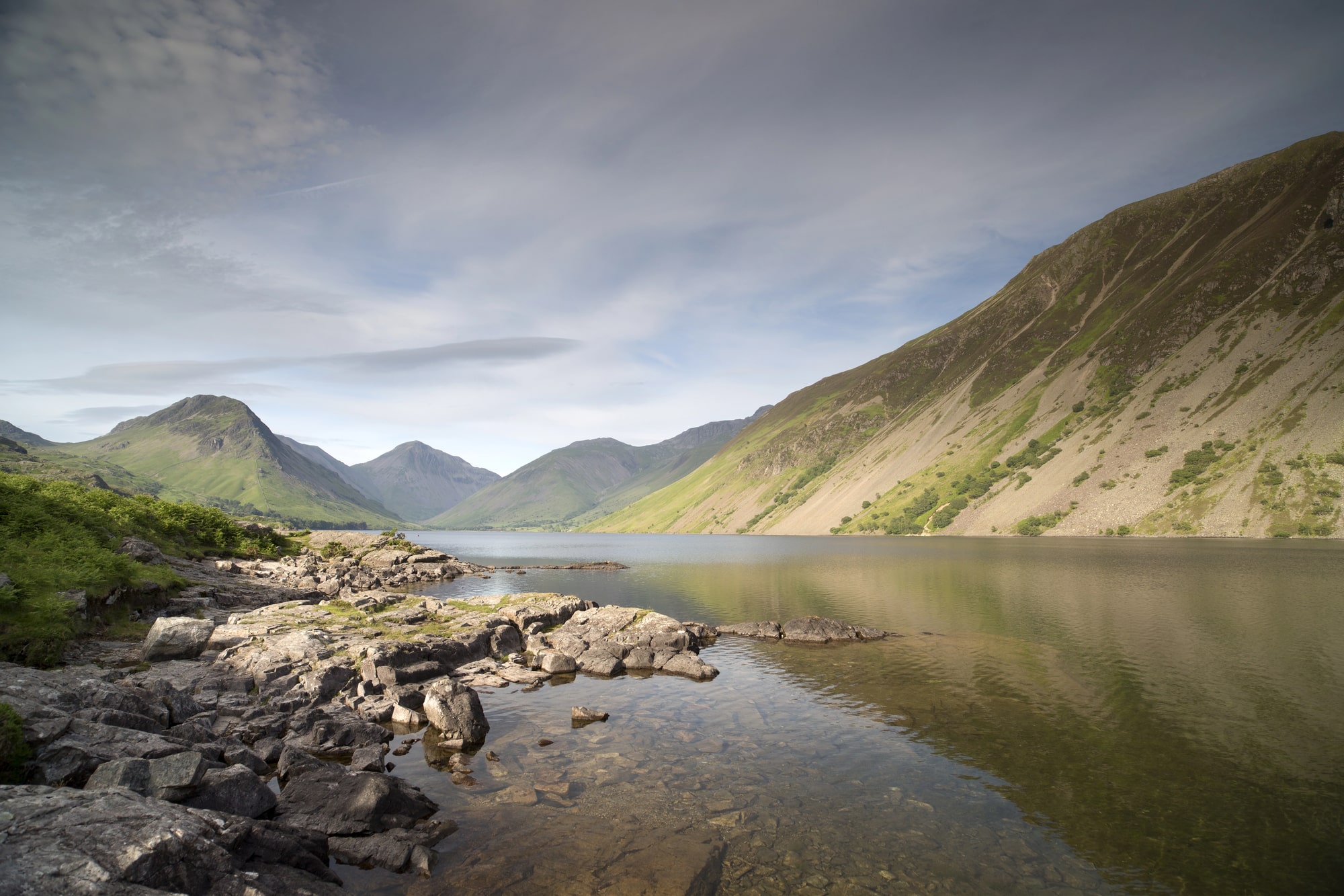Margem rochosa de lago com colinas íngremes sob as nuvens no Lake District